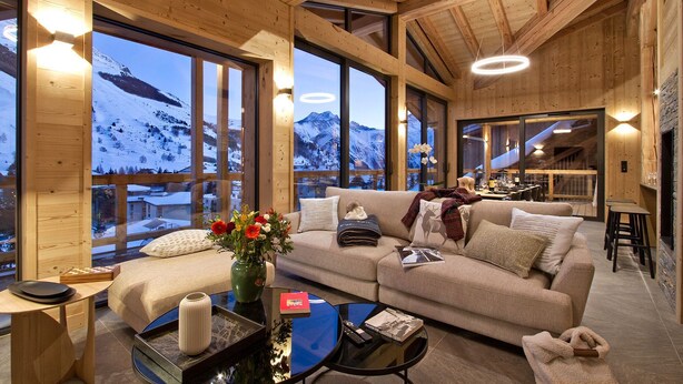 A cozy wooden interior with a sofa, a coffee table, and a vase of flowers. Large windows offer a view of a snowy mountain landscape.
