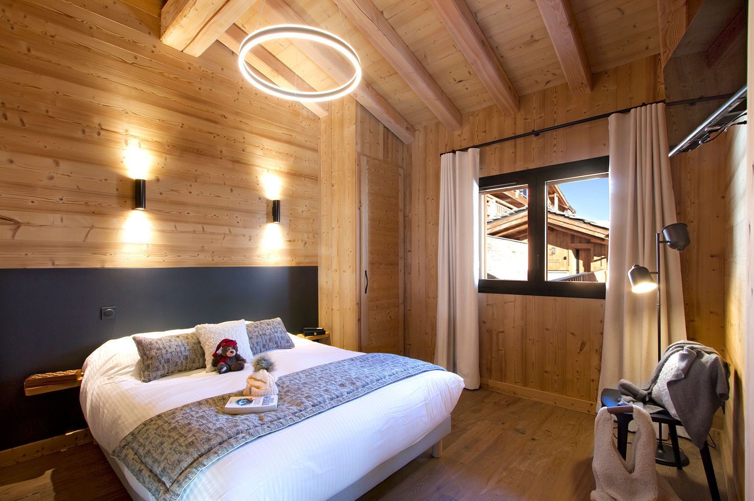 A wooden cabin bedroom with a bed, two wall-mounted lights, a window with curtains, and a view of the cabin's exterior.