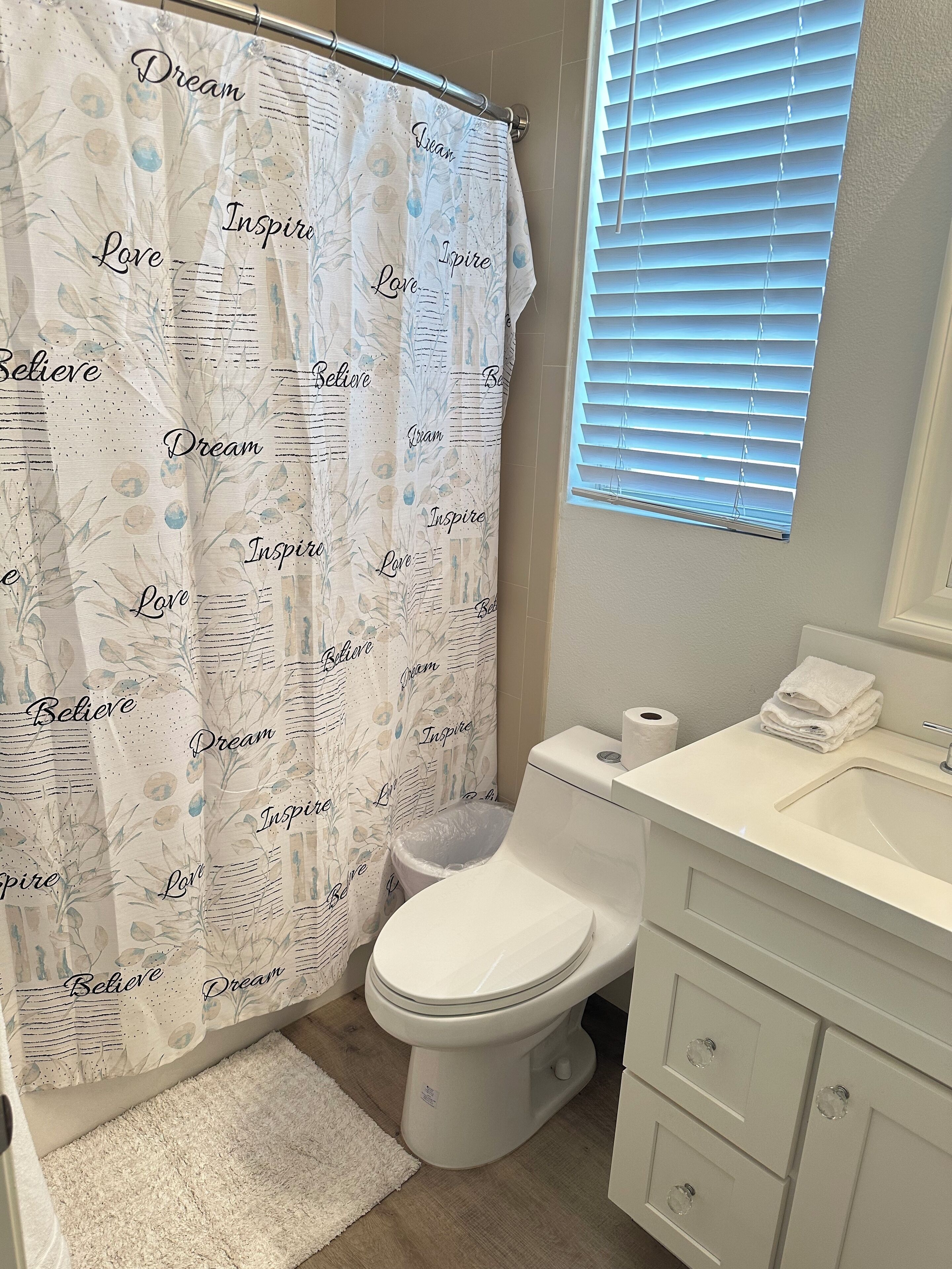 A bathroom with a shower curtain featuring motivational phrases, a toilet, a sink with a mirror, and a window with blinds.