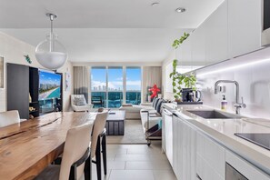 Une cuisine moderne dotée d’un grand îlot central, d’un coin repas avec une table en bois et offrant une vue sur le paysage urbain grâce à de grandes fenêtres.