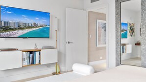 Une chambre d’hôtel moderne avec une grande télévision diffusant une scène de plage, une bibliothèque, une table de chevet et un lit.