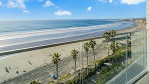 A beachfront view from a balcony overlooking a sandy beach with palm trees and a clear blue sky. - Oceanfront Oasis with Stunning Panoramic Views in Pacific Beach! - Sea It All (San Diego)