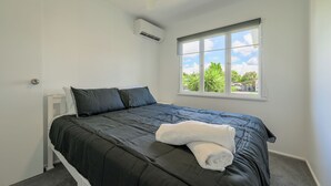 Ein Einzelbett mit dunkler Bettdecke und einem weißen Handtuch, ein Fenster mit Blick auf Grünflächen und ein Deckenventilator.
