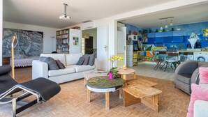 Un salon spacieux avec un canapé, une table basse et un fauteuil de lecture. On aperçoit, à l’arrière-plan, une cuisine aux meubles colorés et un coin repas avec des chaises.