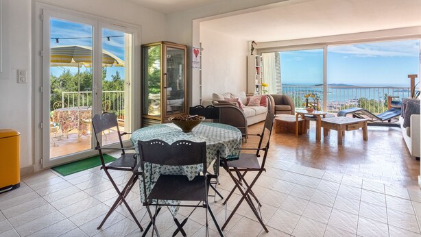 Un vaste salon comprenant un coin repas, de grandes fenêtres offrant une vue sur la mer et un balcon aménagé avec du mobilier d’extérieur.