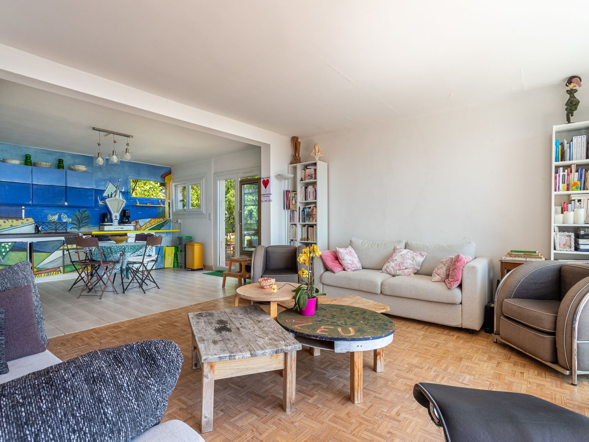 Un vaste espace de vie comprenant un canapé, une table basse et des fauteuils. On peut également apercevoir une cuisine avec des meubles bleus et un coin repas avec des chaises et une table.