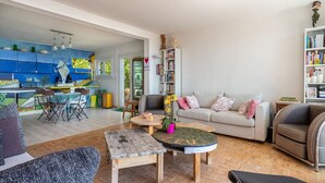 Un vaste espace de vie comprenant un canapé, une table basse et des fauteuils. On peut également apercevoir une cuisine avec des meubles bleus et un coin repas avec des chaises et une table.