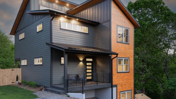 Une maison moderne à deux étages, avec un revêtement extérieur combinant des bois de couleurs foncées et claires, une véranda couverte dotée d’une balustrade et de nombreuses fenêtres.