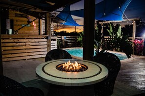 Un'area patio esterna con un braciere circolare, un pavimento in legno e una piscina sullo sfondo.