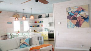 Un salon moderne avec un canapé blanc, une table basse en bois et un ventilateur de plafond. On y trouve également un coin cuisine avec des meubles blancs et un tableau représentant une scène de plage.