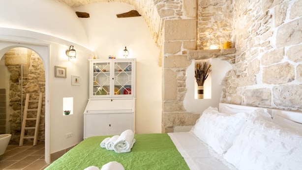 A bedroom with a stone wall, a bed with green bedding, a white cabinet, and a small window with decorative items.