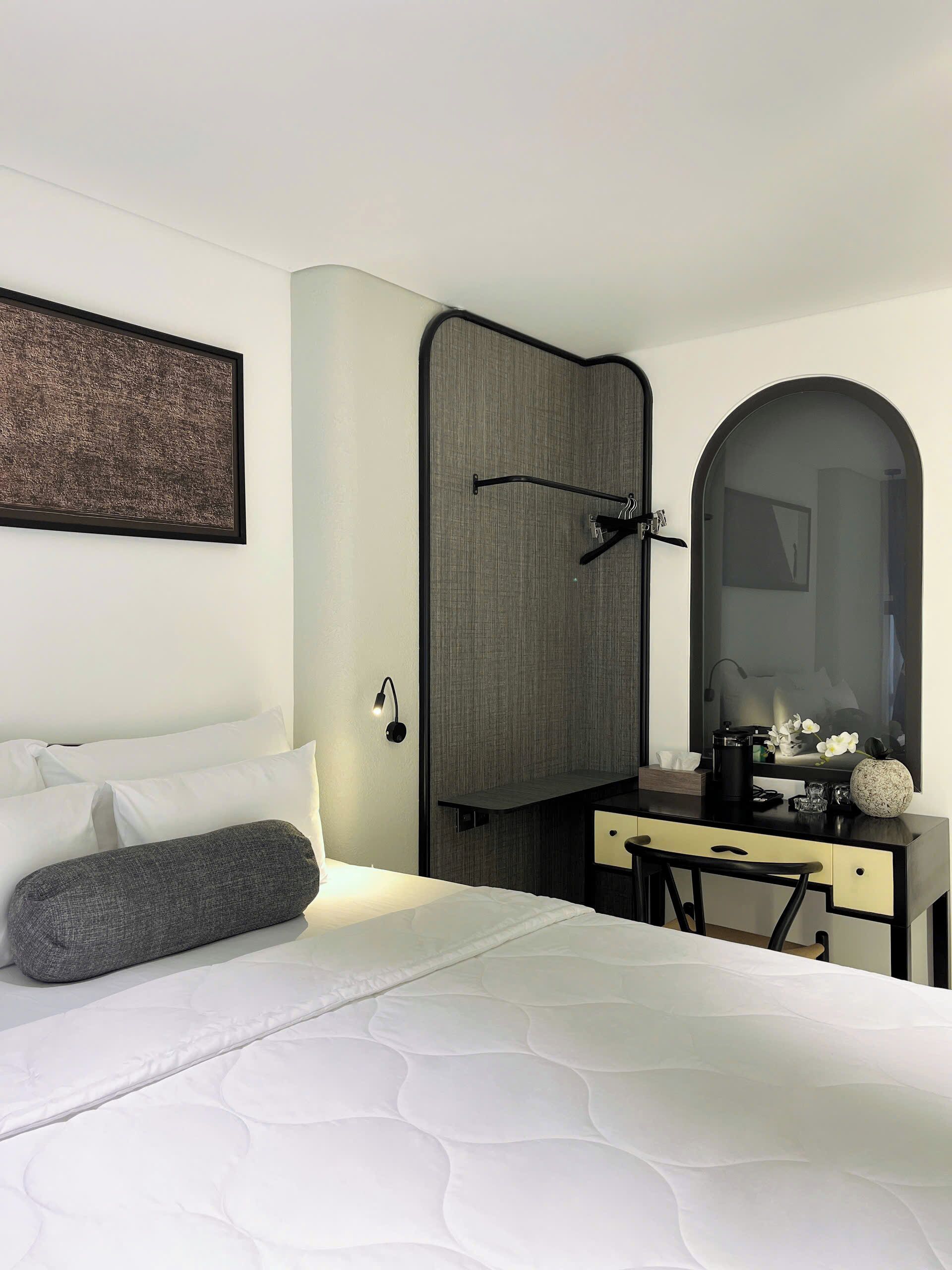 A hotel room with a bed, a desk with a mirror, a wardrobe, and a framed picture on the wall.