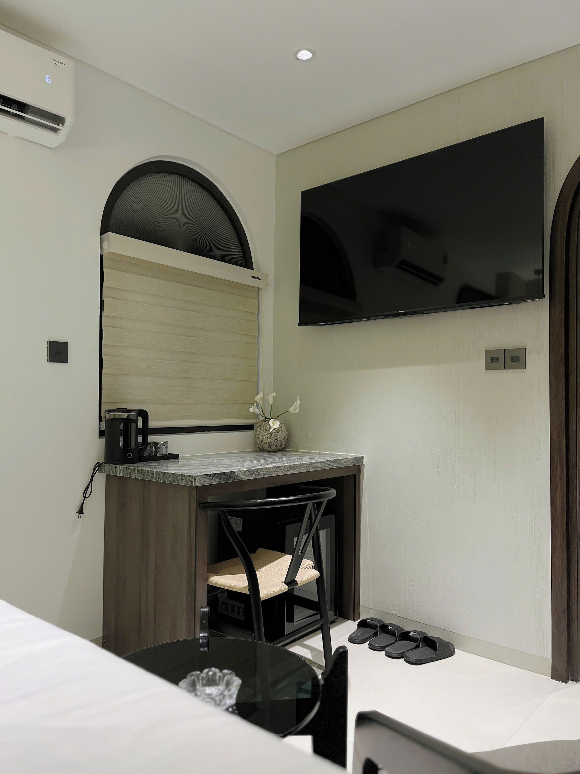 A hotel room with a bed, a desk with a chair, a TV mounted on the wall, and a small table with a vase of flowers.