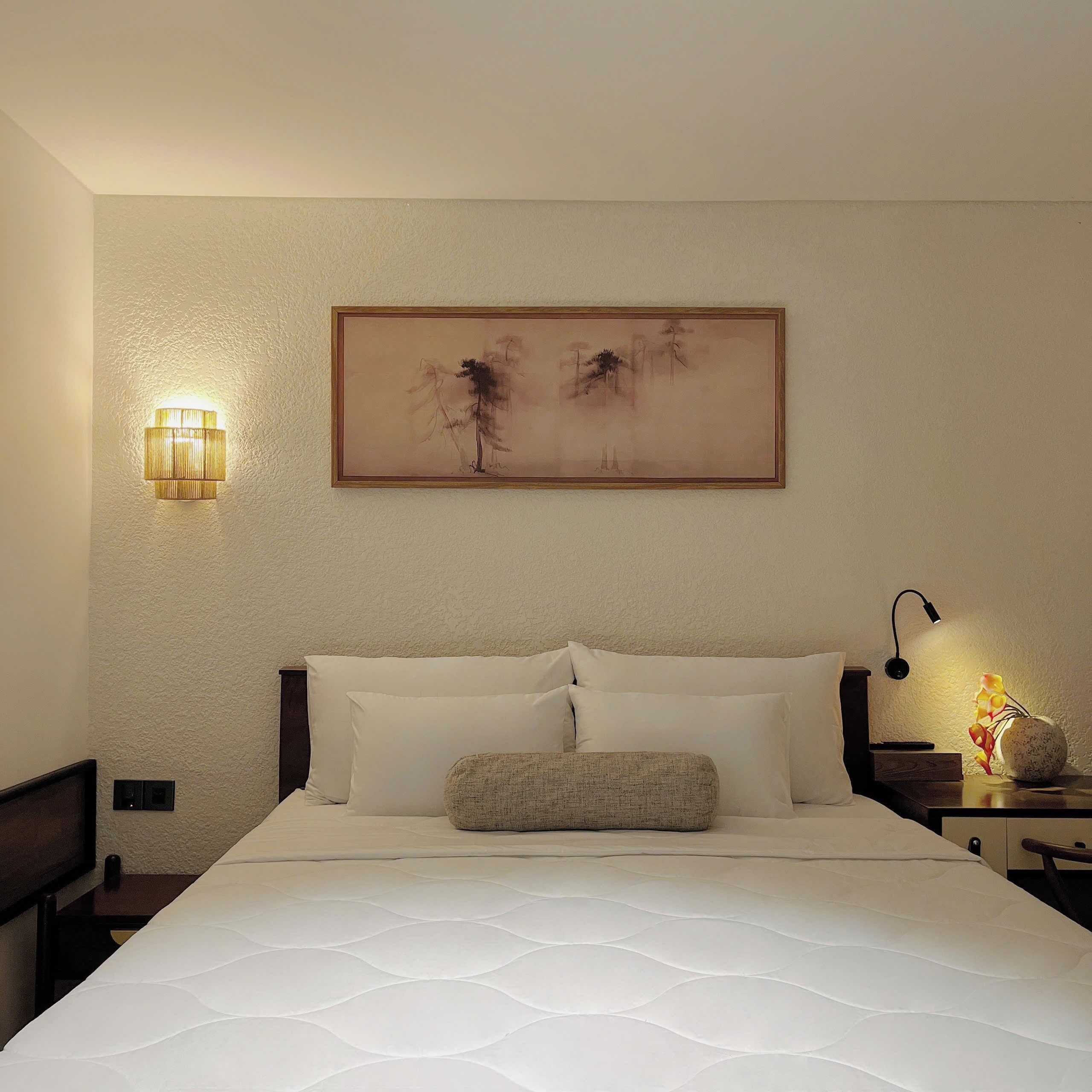 A neatly made bed with white linens, a wall-mounted painting, a bedside lamp, and a small table with a vase.