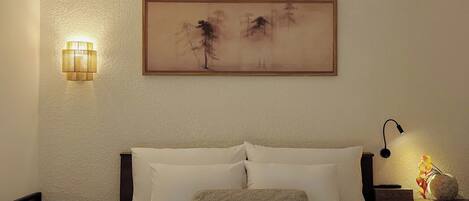 A neatly made bed with white linens, a wall-mounted painting, a bedside lamp, and a small table with a vase.