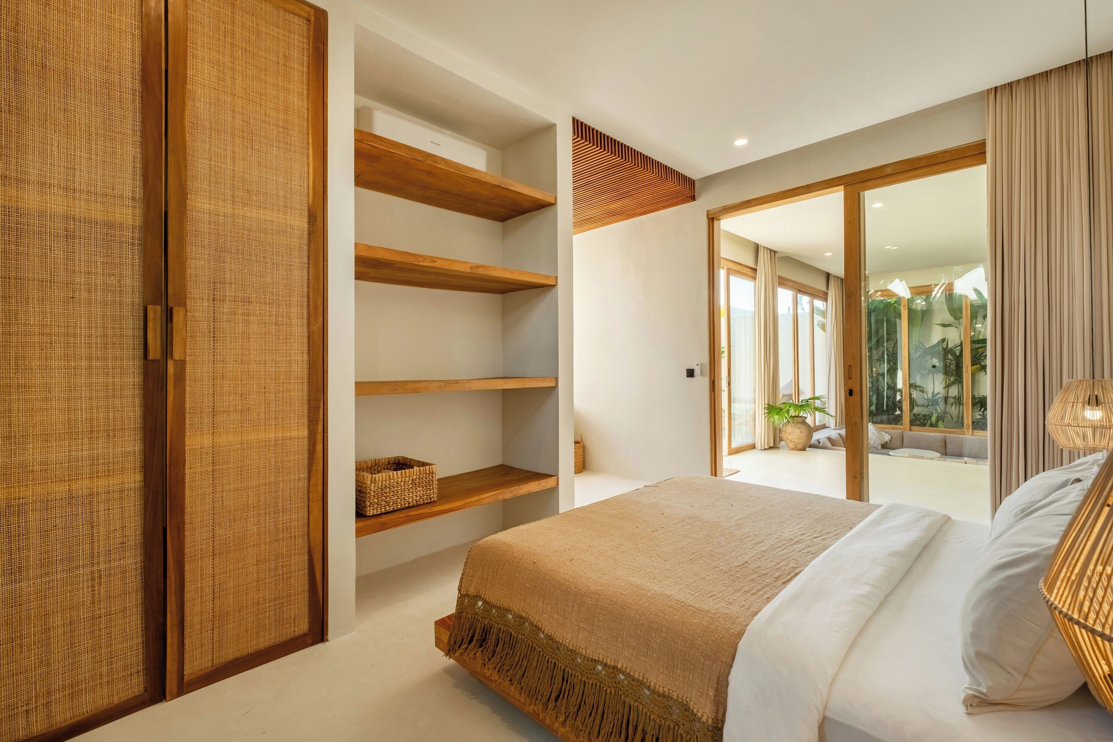 A bedroom with a large bed, wooden shelves, a woven partition, and a view of the outdoors.