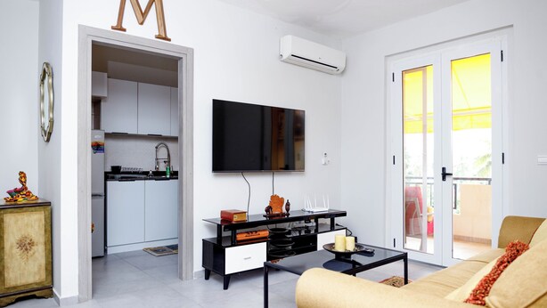 A modern living room with a sofa, coffee table, and TV. There's a kitchen in the background with cabinets and a fridge. - Hope residence Bassam (Modest grand bassam)