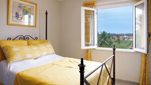 A bed with yellow bedding, a window with curtains, and a view of palm trees and buildings. - Hope residence Bassam (Modest grand bassam)