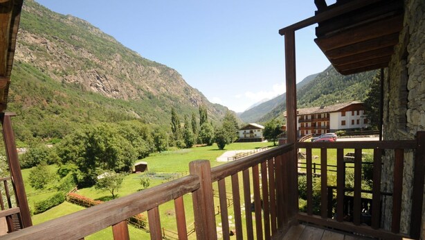 A wooden balcony with a view of a mountainous landscape, a building with a red roof, and a stone wall.