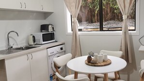 Een compacte keuken met een spoelbak, magnetron en wasmachine. Een ronde tafel met stoelen staat in het midden, en een raam met gordijnen biedt uitzicht op een rotsachtige tuin.