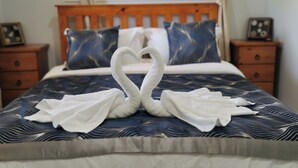 Een bed met een blauw-witte spreideken, twee houten nachtkastjes met ingelijste foto's, en een handdoekopstelling in de vorm van een hart op het bed.