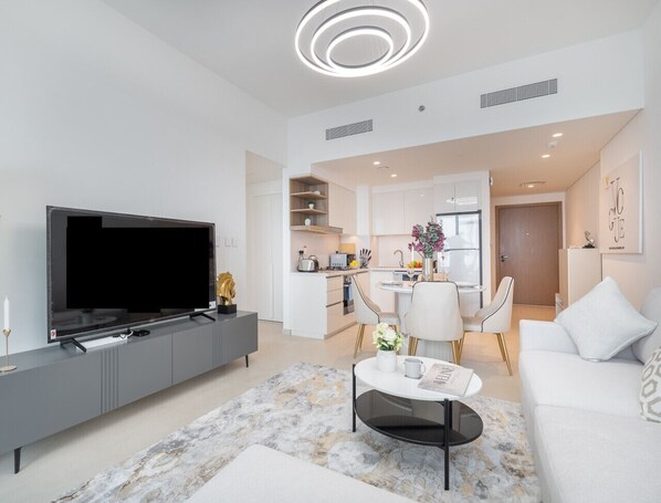 Ein modernes Wohnzimmer mit Flachbildfernseher, grauer Unterhaltungseinheit, weißem Sofa, Couchtisch und einer Küche im Hintergrund.