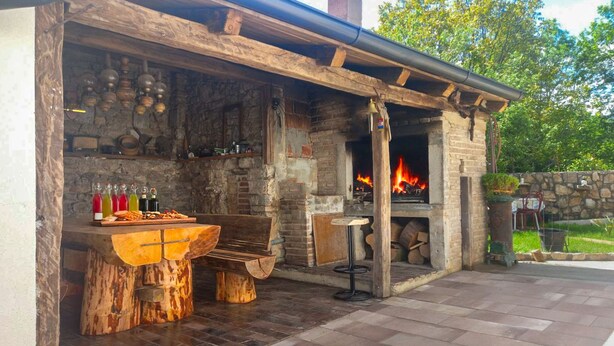 Una chimenea de piedra al aire libre con fuego, una mesa de madera con bebidas y un banco rústico.