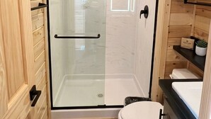 Une salle de bain aux murs lambrissés, dotée d’une douche en verre, de toilettes blanches et d’une étagère où sont rangées les serviettes.