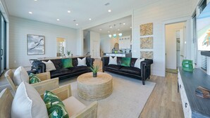 A spacious living room with a black leather sofa, a round woven coffee table, and a dining area with a wooden table and chairs. - Scarlett's Beach Heaven - Luxury Beach House with Rooftop Views and Pool (Miramar Beach)