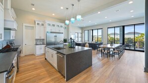 A modern kitchen with a large island, white cabinetry, and stainless steel appliances. There is a dining area with a large table and chairs, and a view of the outdoors through large windows. - Scarlett's Beach Heaven - Luxury Beach House with Rooftop Views and Pool (Miramar Beach)