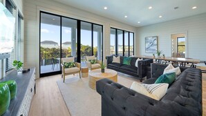 A spacious living area with a large black sofa, a dining table, and a view of the outdoors through large sliding glass doors. - Scarlett's Beach Heaven - Luxury Beach House with Rooftop Views and Pool (Miramar Beach)