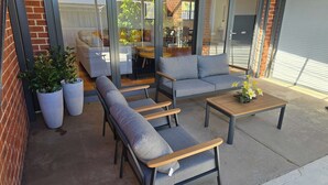 Een terras met een bank, stoelen en een bijzettafeltje. Er staan potplanten en een glazen deur naar een binnenruimte.