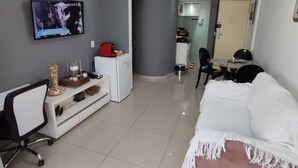 A modern living room with a TV, a white sofa, a pink ottoman, a desk with a chair, and a kitchen area in the background. - ACOMODAÇÕES RG APARTAMENTOS SÃO PAULO SP (São Paulo)