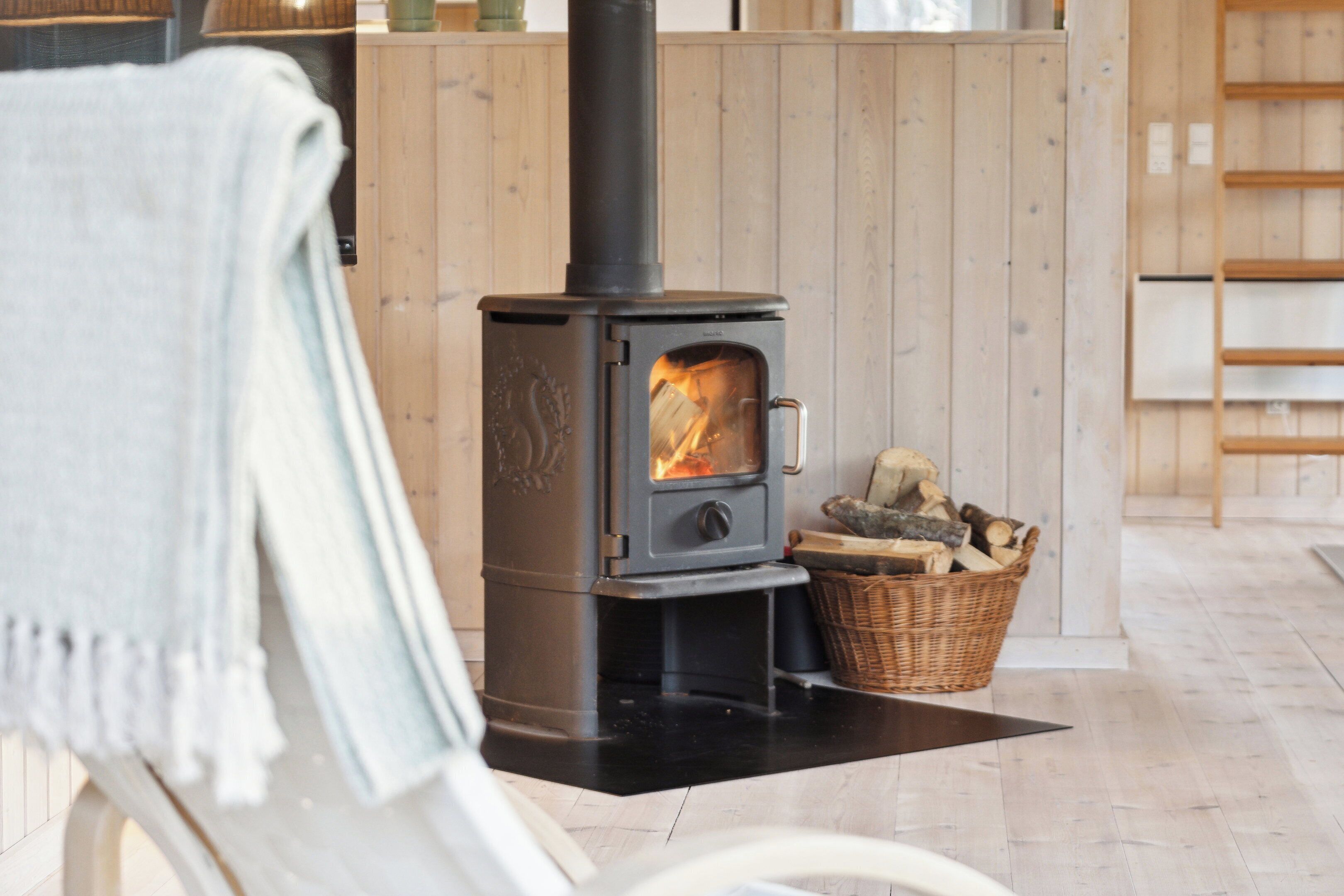 A room with a wood-burning stove, a wicker basket with logs, and a white chair with a grey throw.