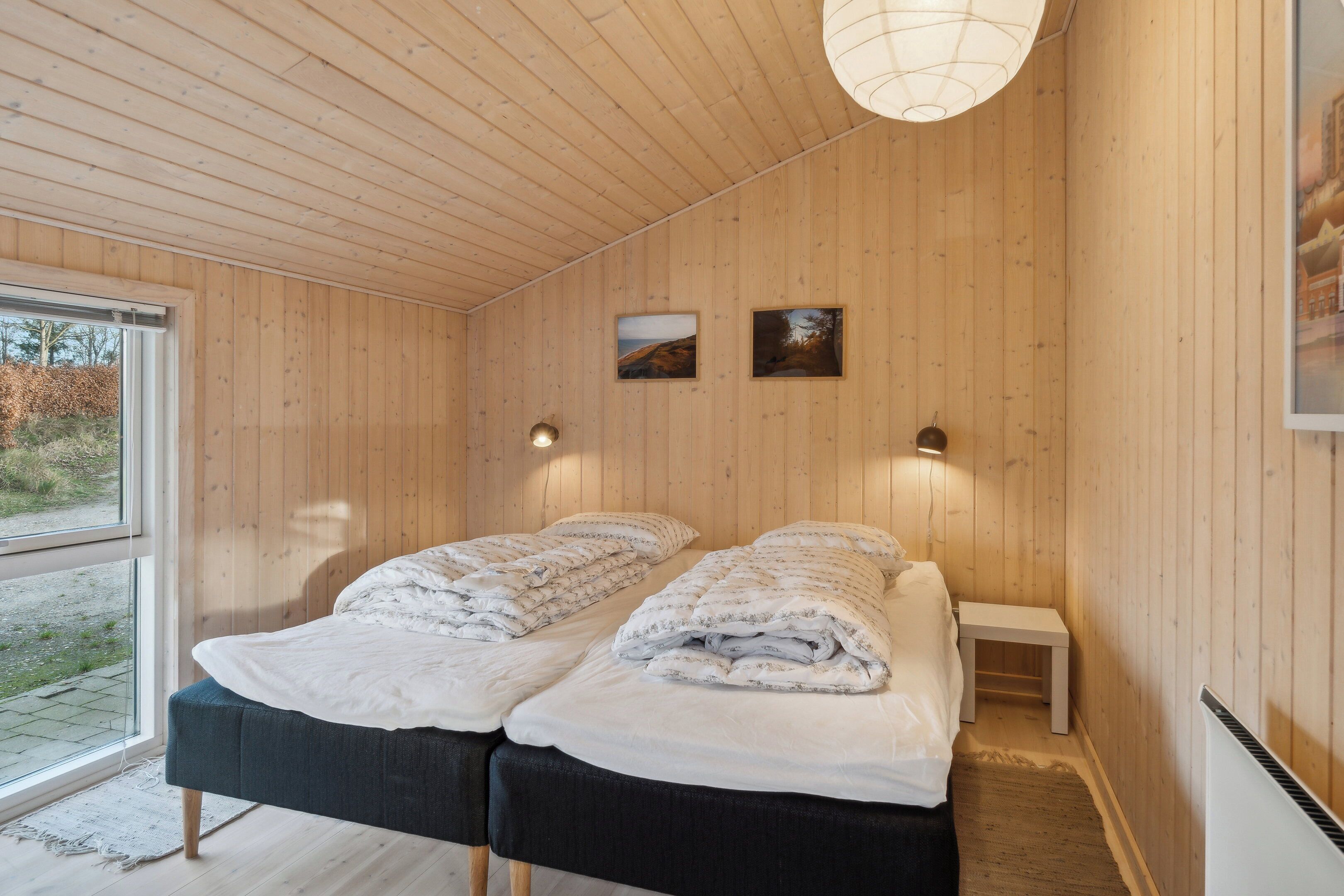 Ein Zimmer mit Holzwänden, zwei Betten mit weißer Bettwäsche, ein kleiner Nachttisch und ein Fenster mit Blick auf Grünflächen.