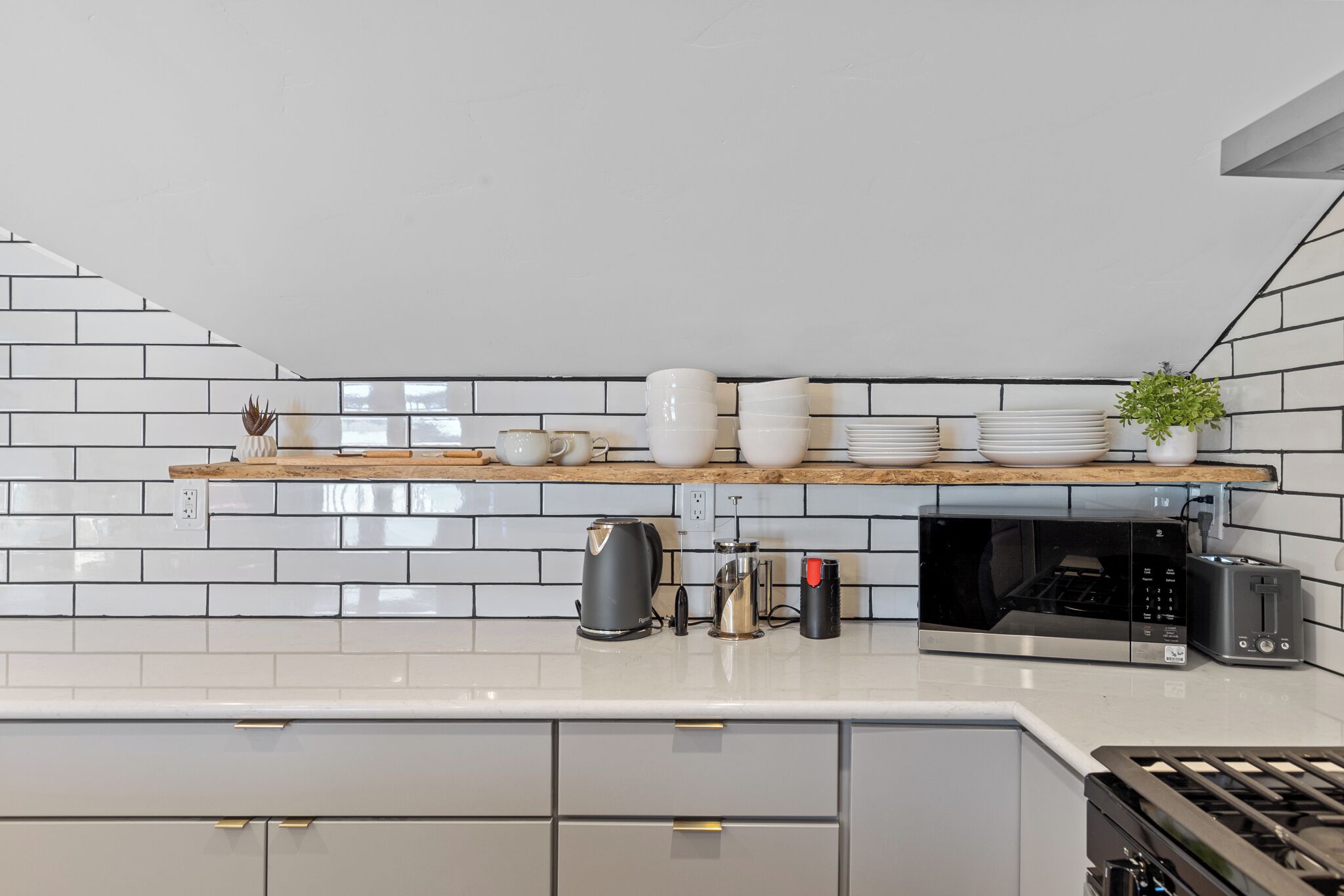 A modern kitchen with white subway tiles, a wooden shelf with dishes, a coffee maker, a toaster, and a microwave.