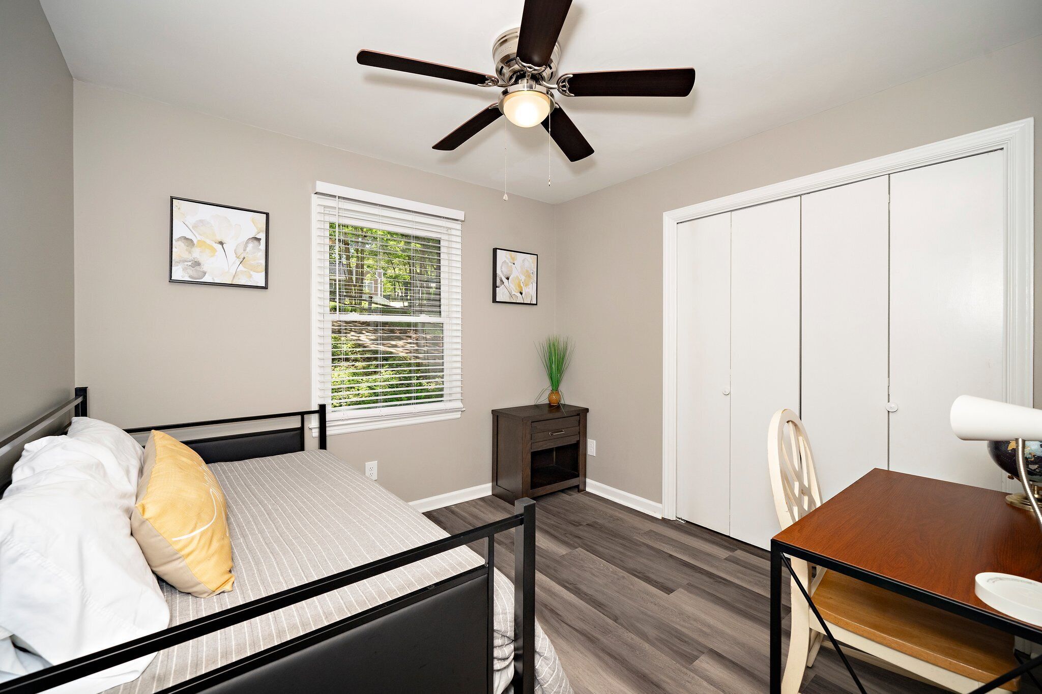 A bedroom with a bed, a desk, a chair, a wardrobe, a ceiling fan, and a window with blinds.