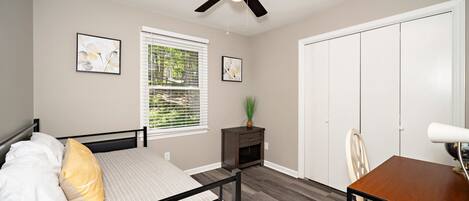 A bedroom with a bed, a desk, a chair, a wardrobe, a ceiling fan, and a window with blinds.