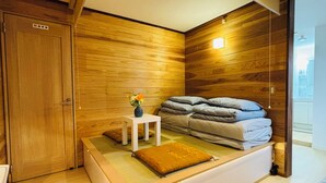 A modern bedroom with a wooden wall, a bed with grey bedding, a small white table with a vase, and a wooden door.