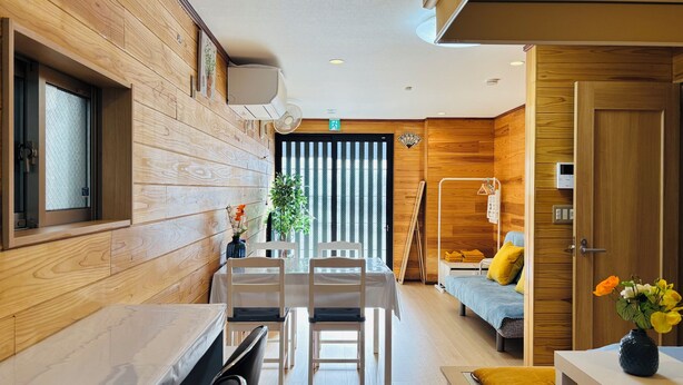 A modern hotel room with wooden paneling, a dining area with a table and chairs, a bed with yellow pillows, and a small table with a vase of flowers.