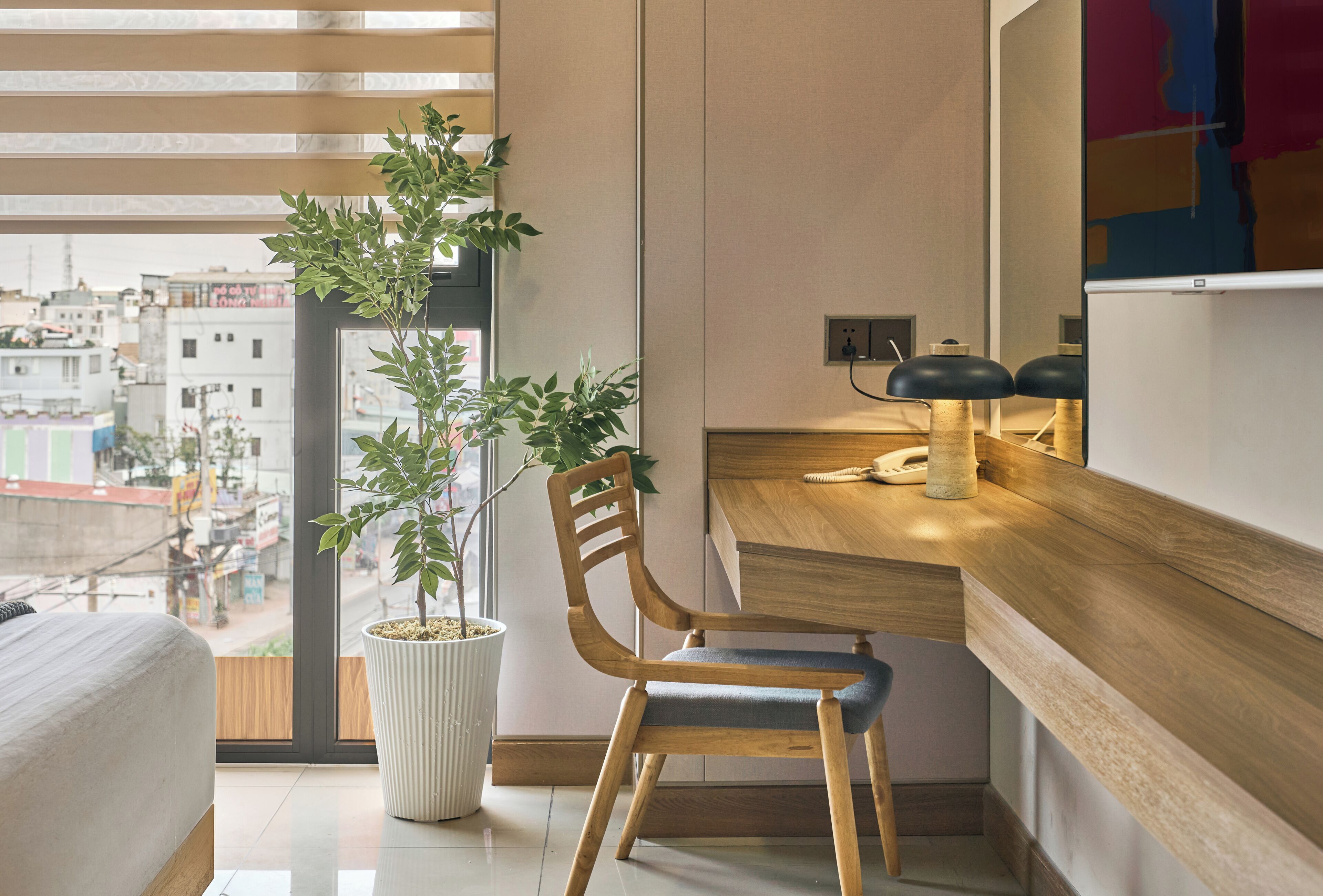 Une chambre d’hôtel moderne avec un bureau en bois, une chaise, une plante en pot et une vue sur la ville.