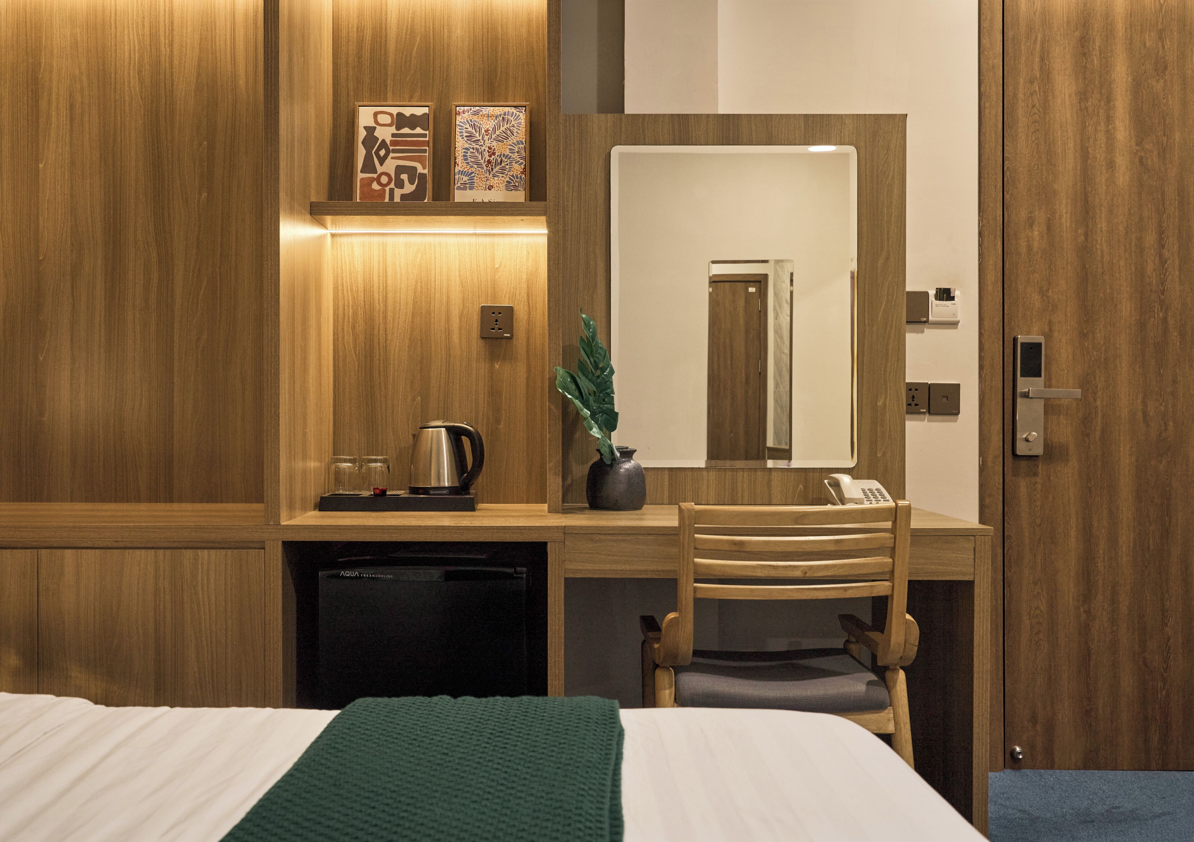 A hotel room with a wooden desk, a bed with a green blanket, a potted plant, a mirror, and a telephone.