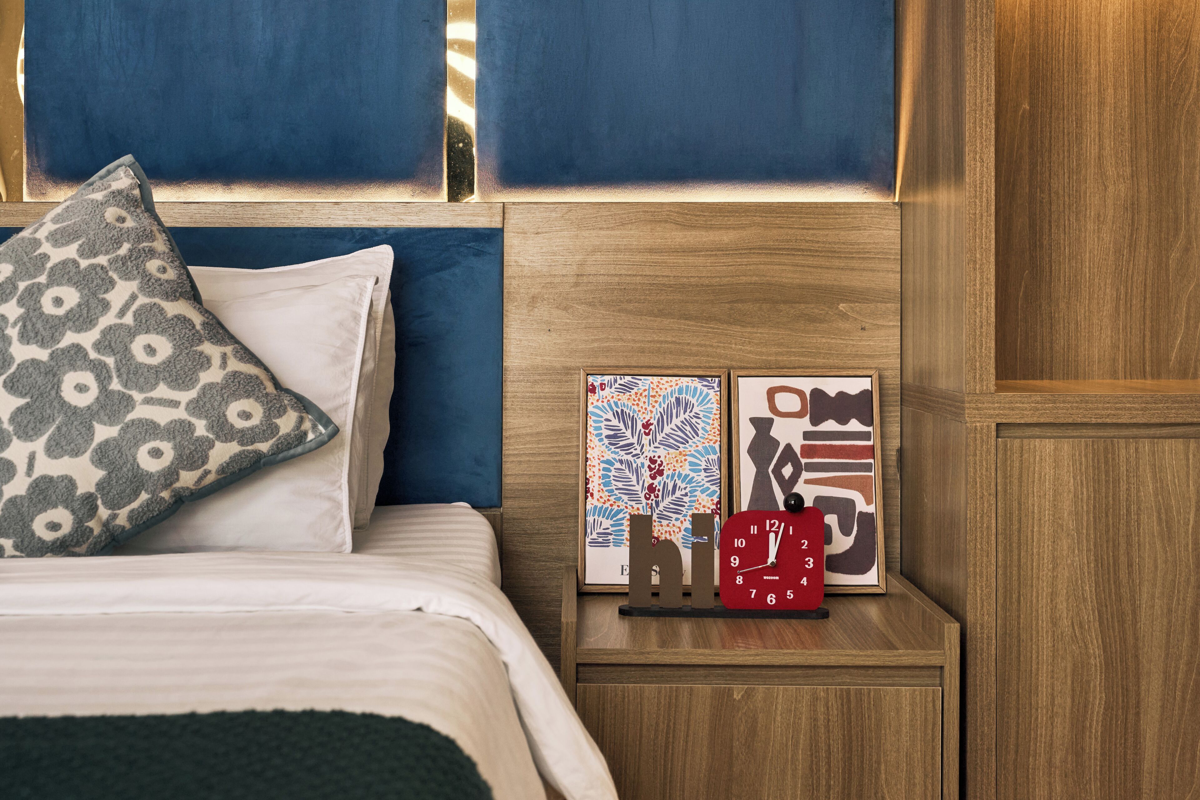 Una cama bien hecha con un cojín estampado, una mesita de noche con un reloj y una obra de arte enmarcada, y un cabecero de madera.
