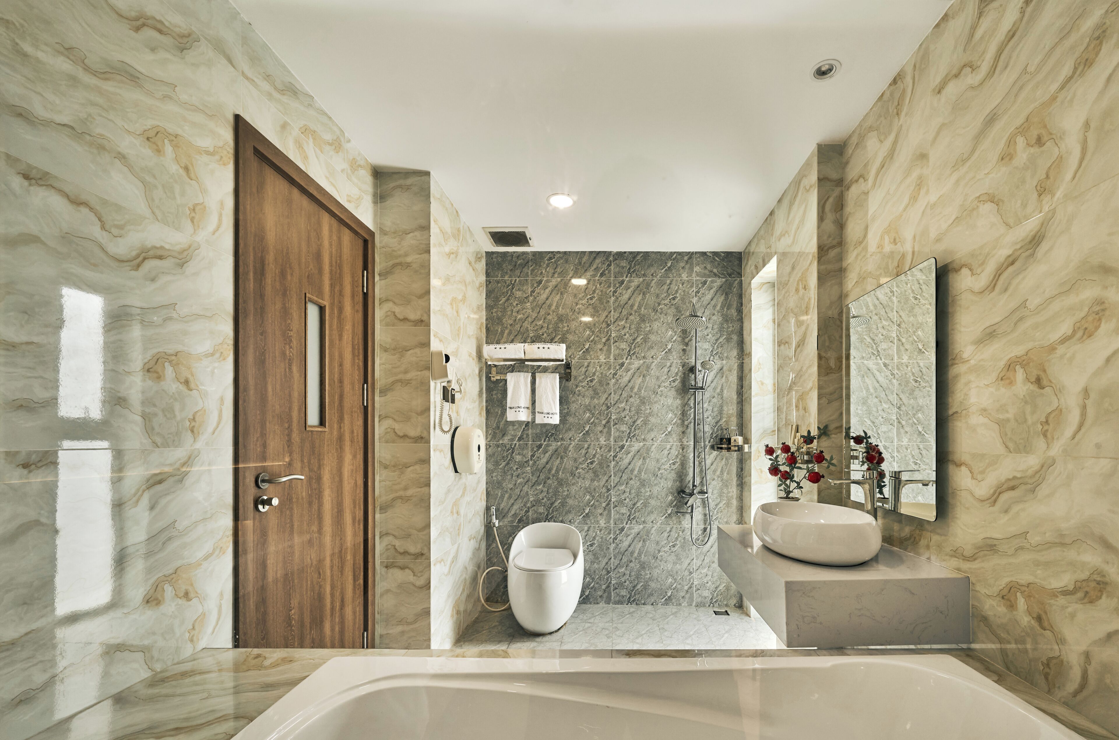 Un baño moderno con pared de mármol, puerta de madera, inodoro blanco, ducha, lavamanos y un espejo grande.
