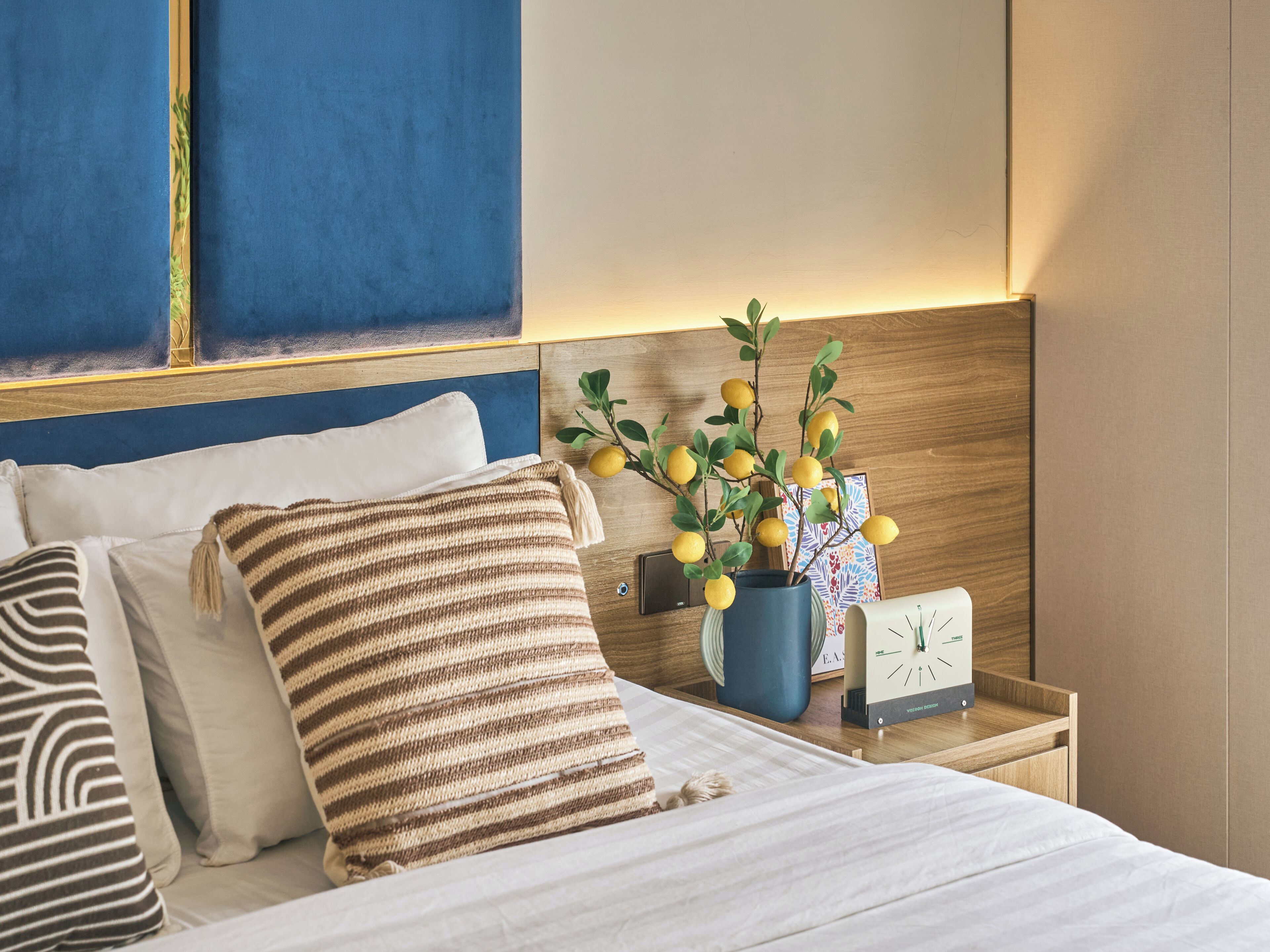 A neatly made bed with a striped blanket and pillows, a bedside table with a clock and a vase of flowers, and a blue headboard.