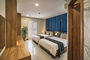 Habitación de hotel con dos camas, un ventanal con persianas, una planta en maceta y una pared de acento azul.