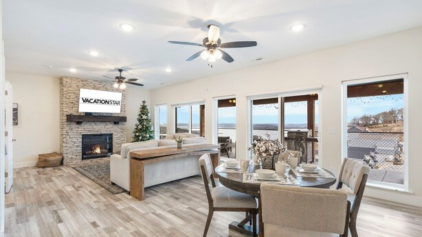 A spacious living room with a fireplace, a stone accent wall, a ceiling fan, and large windows offering a view of the outdoors.