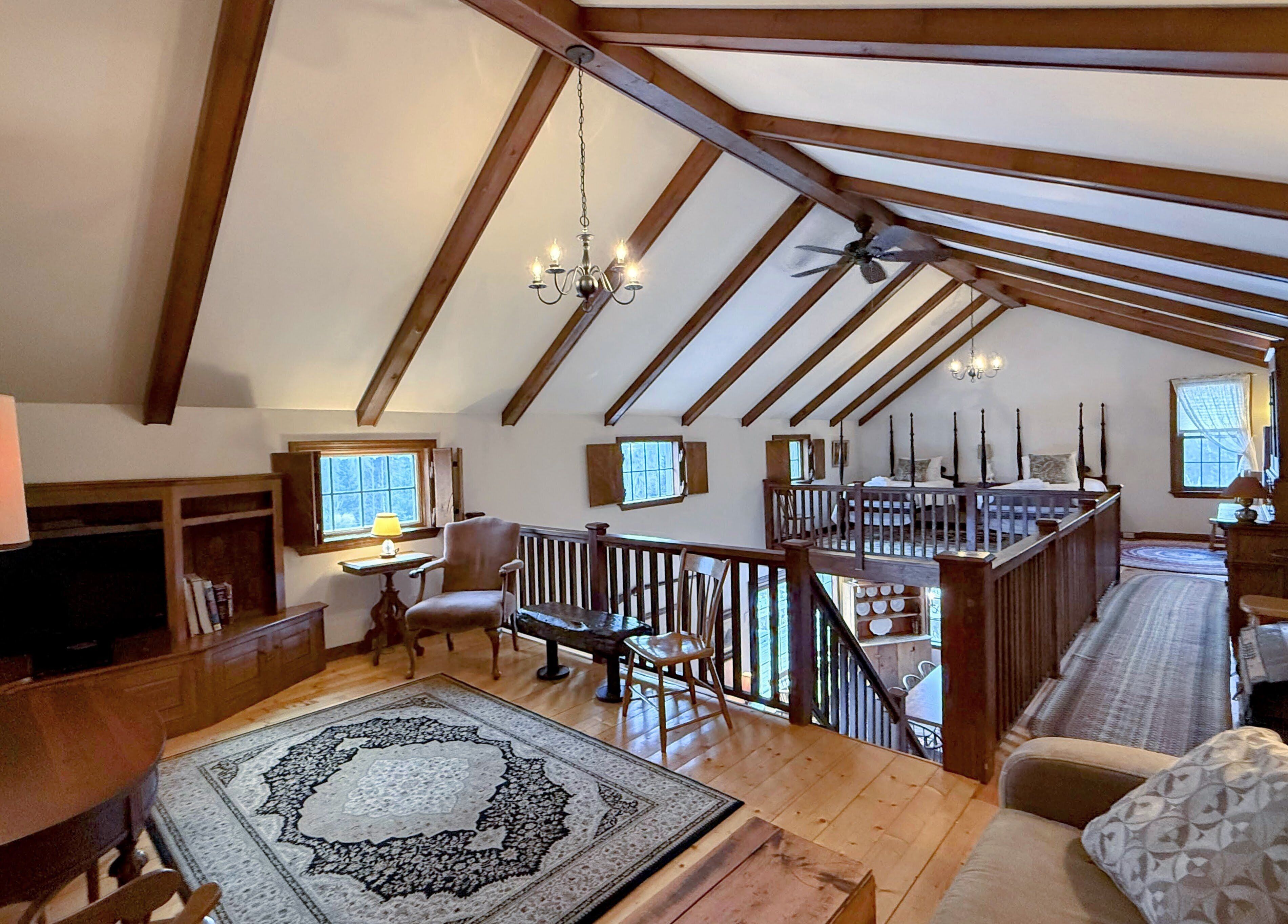 A spacious loft bedroom with a high ceiling, wooden beams, a large bed, a desk, and a staircase leading to an upper level.