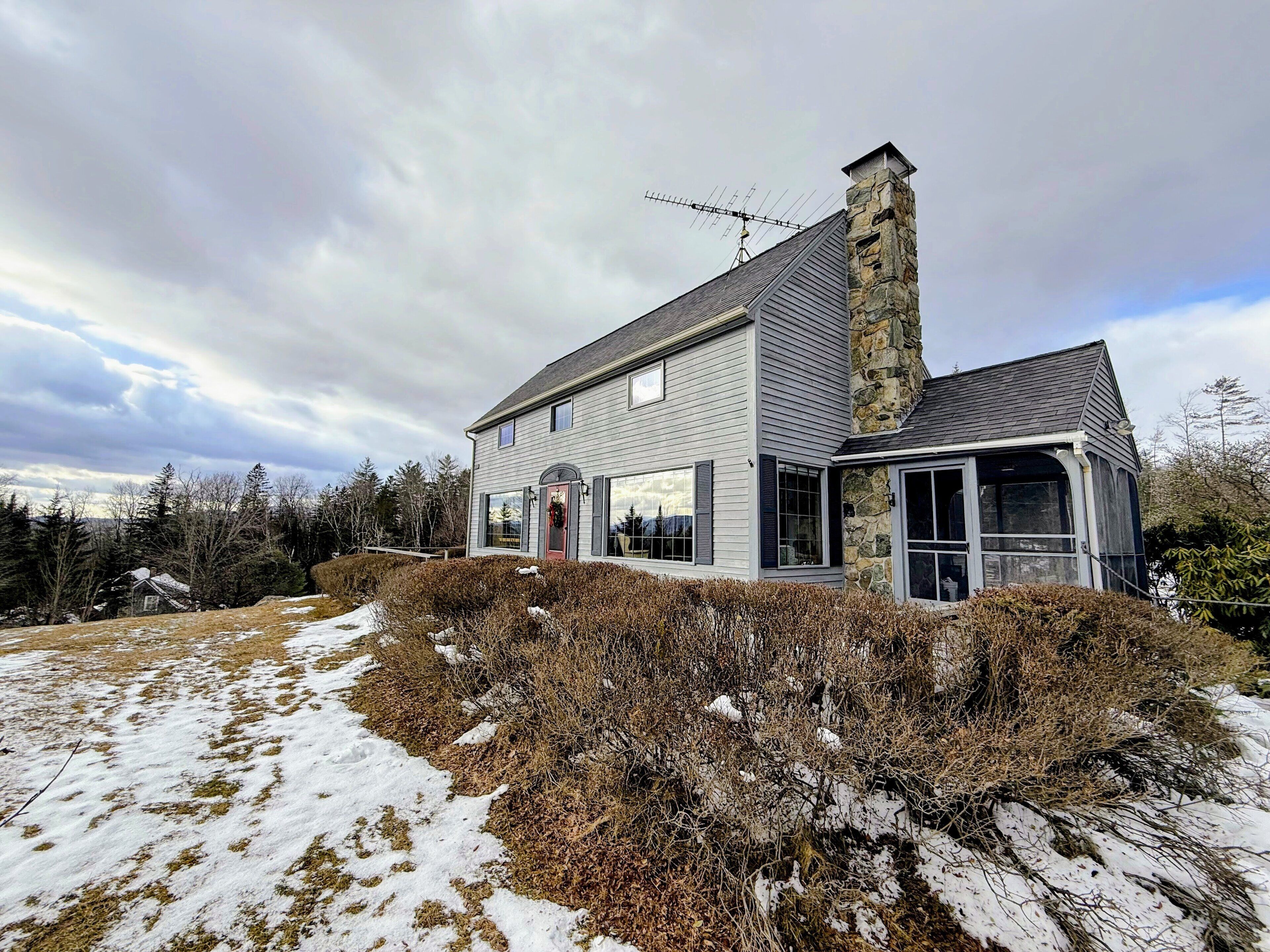 A two-story house with a stone chimney, large windows, and a snow-covered landscape.