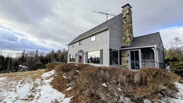 A two-story house with a stone chimney, large windows, and a snow-covered landscape.